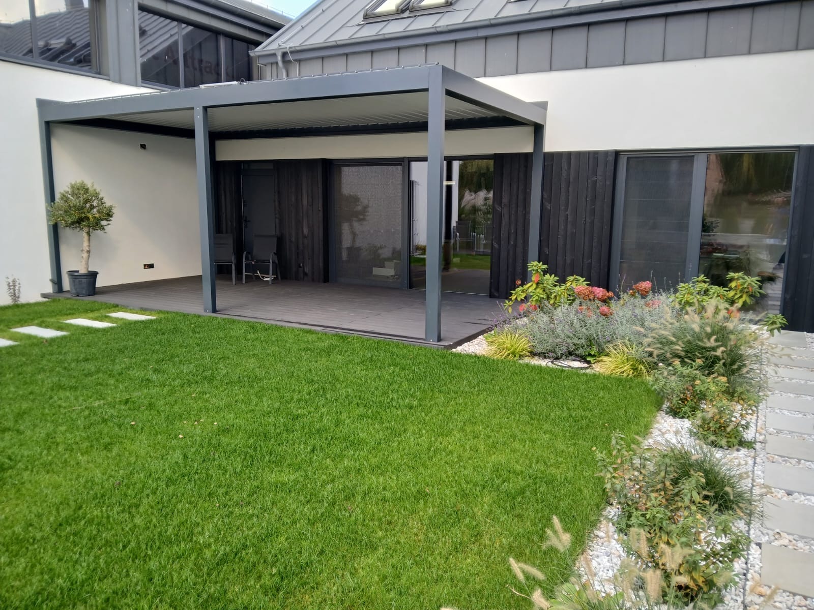 Nowoczesna pergola tarasowa z szarymi słupami i zadaszeniem, widok z zewnątrz na zielony trawnik i rabaty kwiatowe. Elegancki design, drewniane wykończenie podłogi.