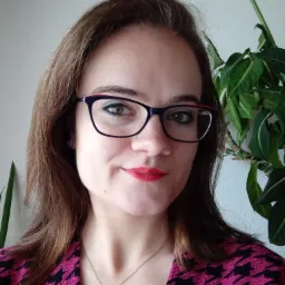 Portret uśmiechniętej kobiety w okularach z czerwoną szminką, w różowo-czarnej marynarce w pepitkę i srebrnym naszyjnikiem, z rośliną w tle.