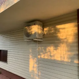 Zewnętrzna jednostka klimatyzacji Gree zamontowana na elewacji domu z sidingu, widoczne rury i przewody, ciepłe światło zachodzącego słońca