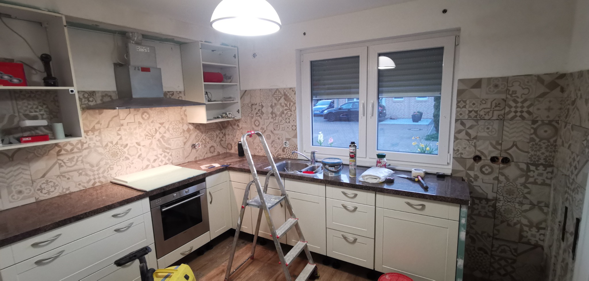 Kuchnia w trakcie prac: białe szafki z ciemnym blatem, wzorzyste płytki na ścianie, drabina, narzędzia i materiały budowlane widoczne na blacie, brakujące fronty szafek górnych.