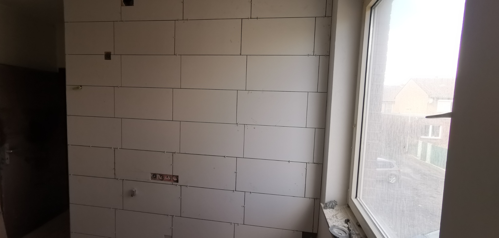 Ściana wyłożona płytami kartonowo-gipsowymi, widoczne otwory na gniazdka elektryczne i okno z widokiem na zewnątrz.