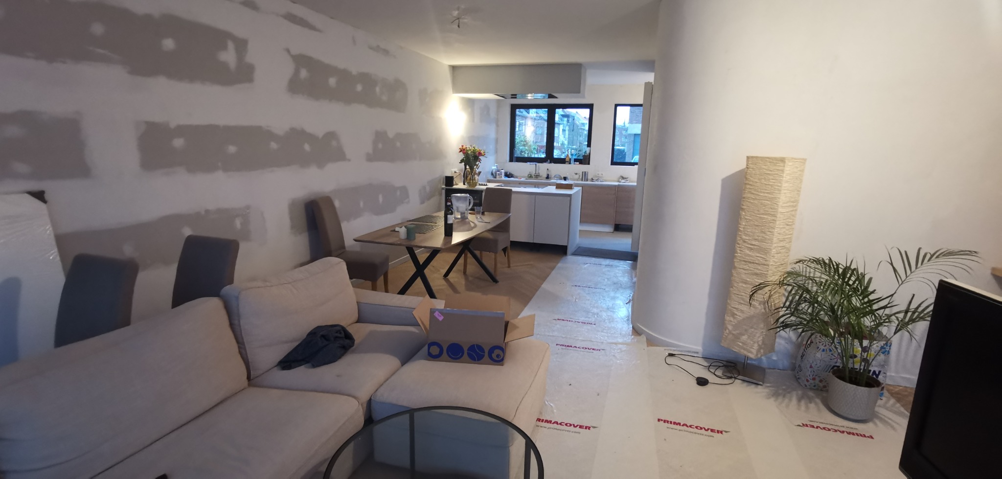 Pokój w trakcie prac przygotowawczych do wykończenia, ściany z widocznymi pasami po nakładaniu gładzi, podłoga zabezpieczona folią, widoczny fragment salonu z sofą i jadalni z stołem i krzesłami...