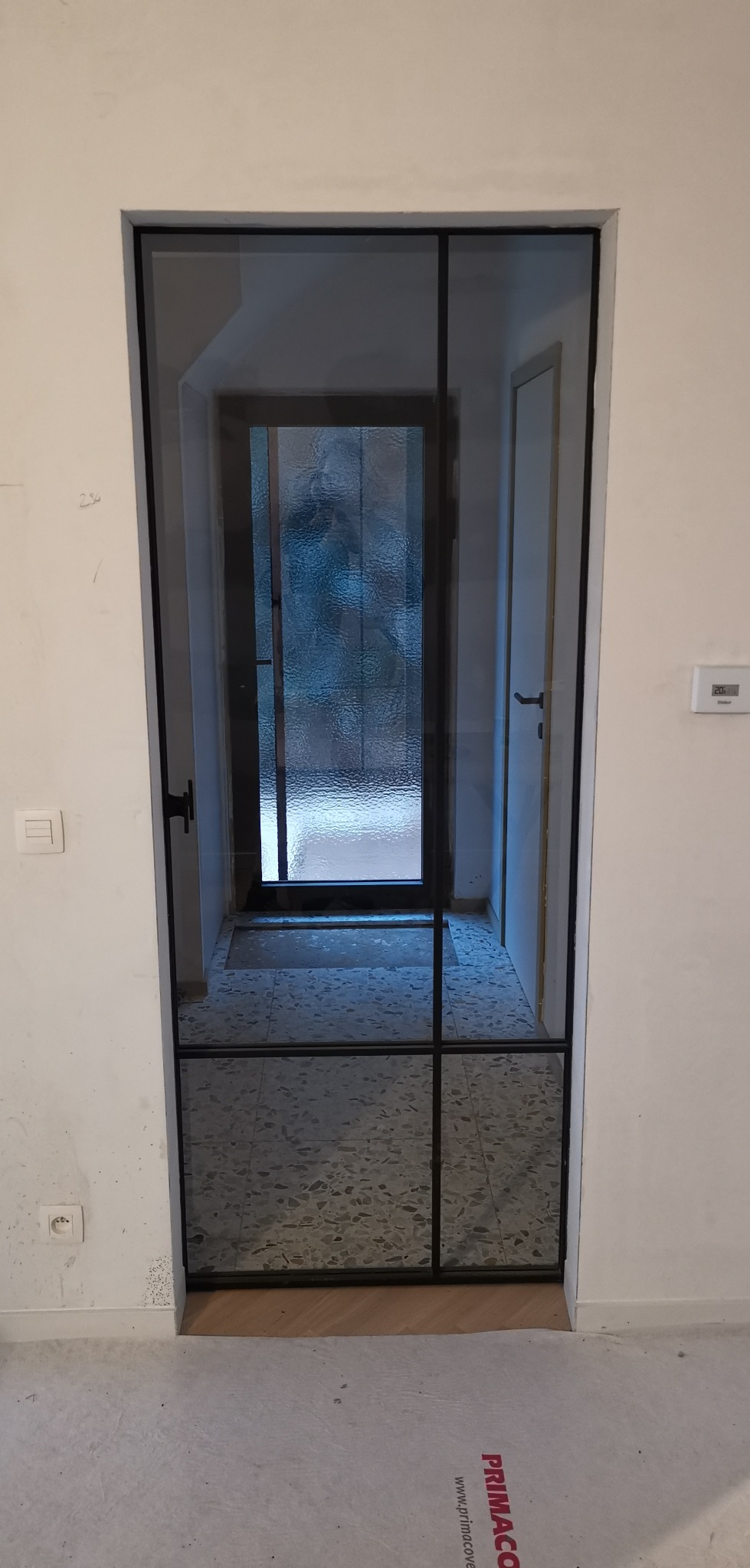 Nowoczesne, czarne drzwi wewnętrzne z przeszkleniami, oddzielające korytarz z terazzo od dalszej części domu, zabezpieczone folią ochronną na podłodze.