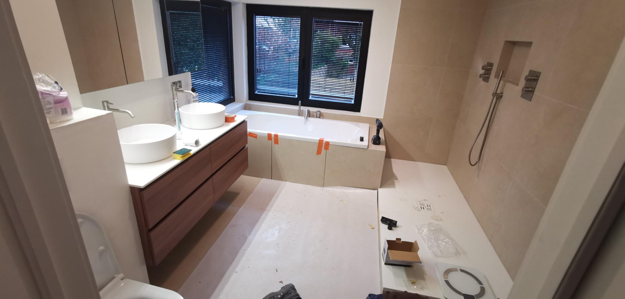 Łazienka w trakcie wykańczania w Ełku: widoczne dwie białe umywalki na drewnianej szafce, wanna obudowana płytami, prysznic w ścianie, narzędzia i materiały budowlane na podłodze.