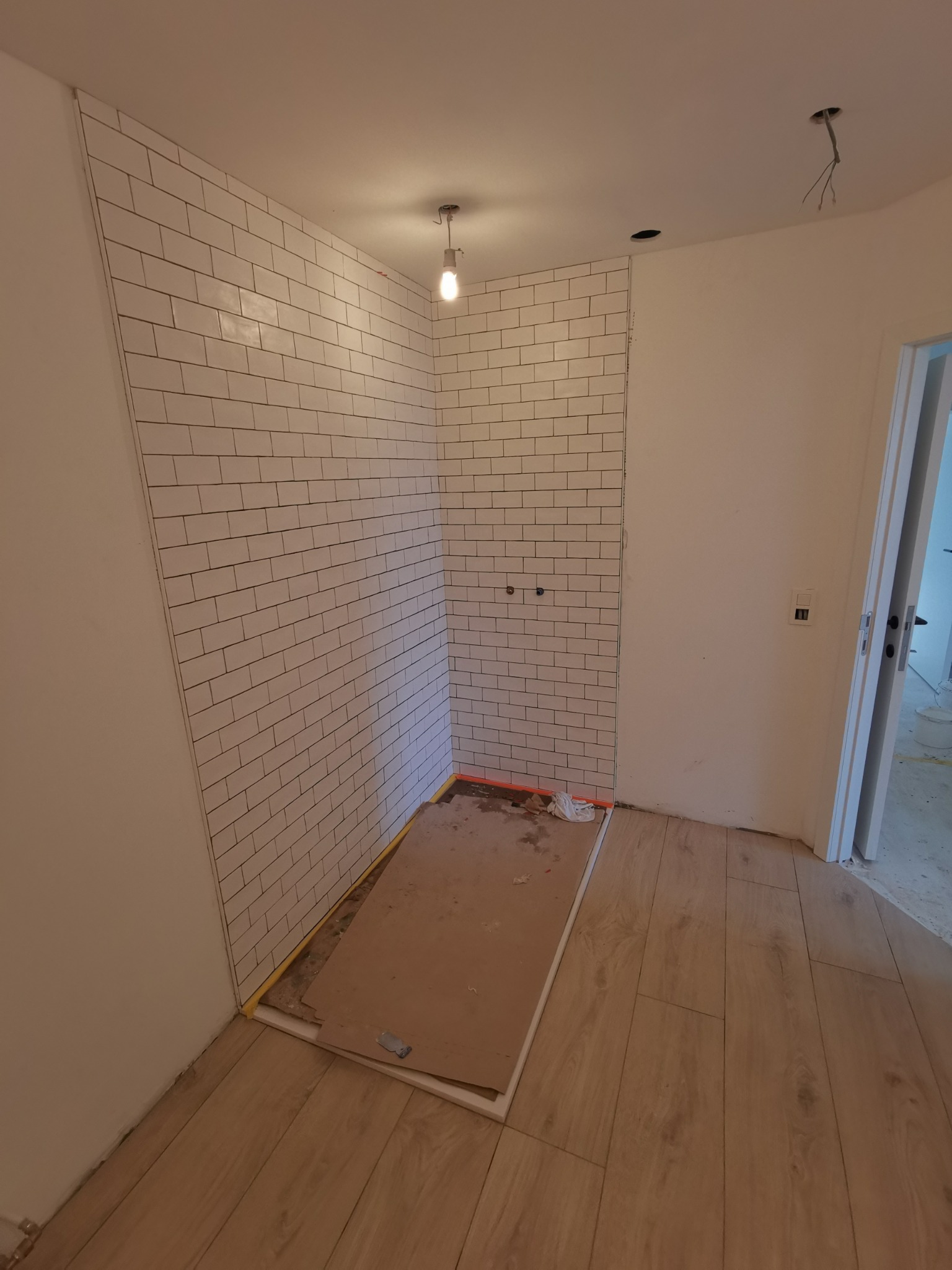 Ściana wyłożona białymi, prostokątnymi płytkami w trakcie prac, widoczne niedokończone fugi i wystające rury, podłoga zabezpieczona kartonem, w tle drewnopodobne panele i otwarte drzwi.