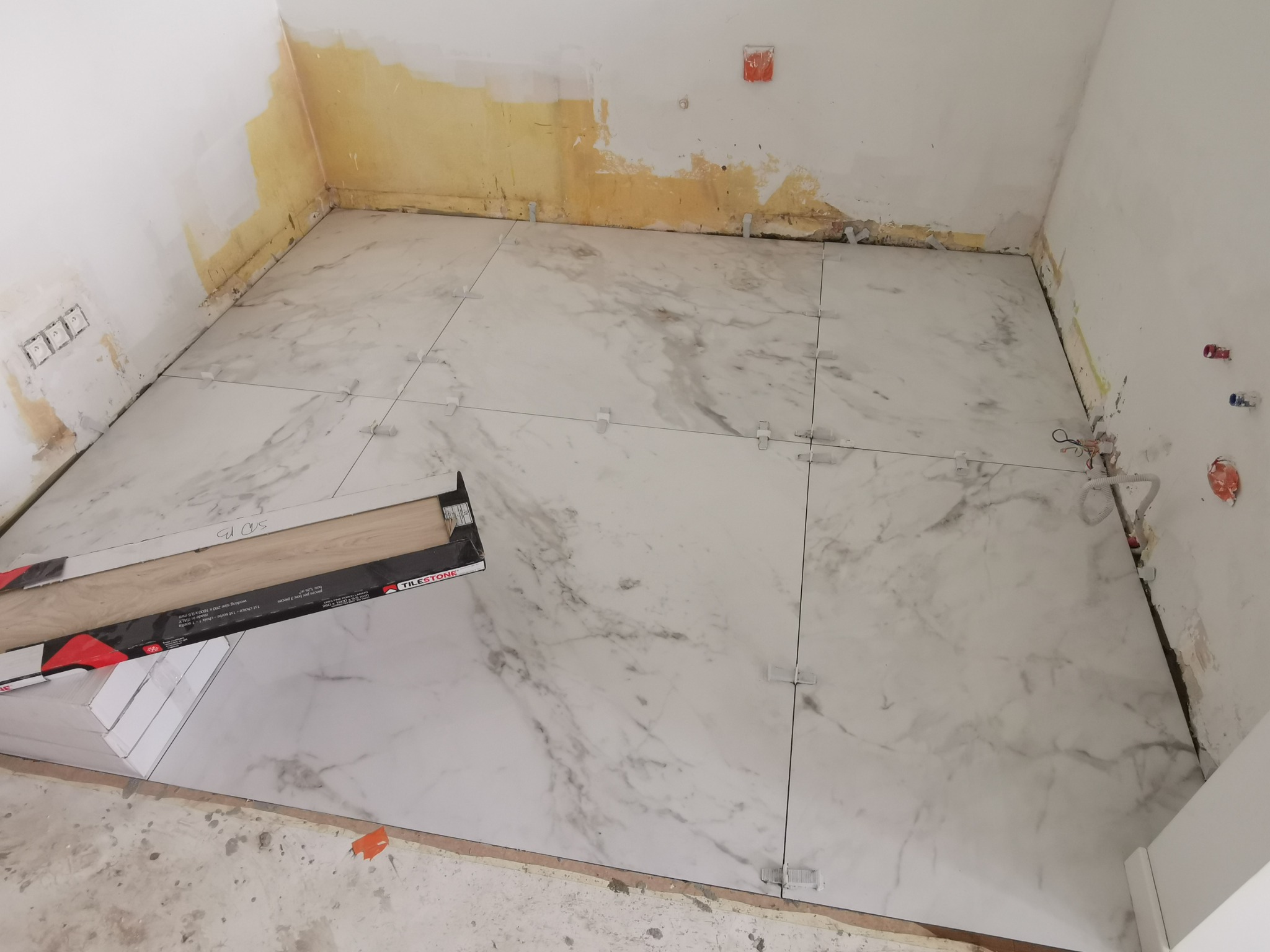 Układanie białych płytek imitujących marmur na podłodze w trakcie prac wykończeniowych, widoczne krzyżyki dystansowe i paczka płytek w kartonie firmy TILSTONE.
