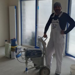 Usługi remontowe Krzysztof Dąbrowski - Malarz w białym kombinezonie Flűgger farby, z maszyną Graco Ultra Max II 795, worki z farbą w tle, okna zabezpieczone folią malarską. Przygotowanie do malowania.