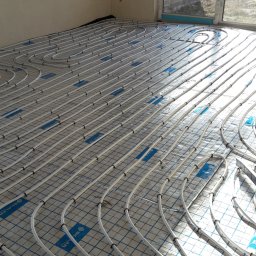 Usługi remontowe Krzysztof Dąbrowski - Białe rury ogrzewania podłogowego ułożone równolegle na niebieskiej siatce w jasnym pomieszczeniu z dużym oknem. Widoczny fragment ściany i surowy stan wykończenia.