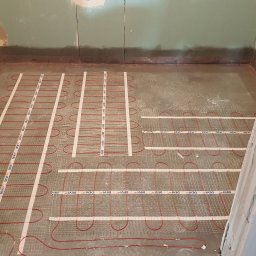 Usługi remontowe Krzysztof Dąbrowski - Montaż ogrzewania podłogowego w łazience, widoczne kable grzewcze marki Devi na siatce, przygotowane do zalania wylewką. Ściany surowe, w tle płyta karton-gips.