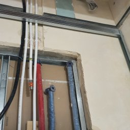 Usługi remontowe Krzysztof Dąbrowski - Instalacja wodna w trakcie montażu: białe, czerwone i niebieskie rury prowadzone wzdłuż ściany z profilem metalowym. Widoczny bojler i elementy mocujące.