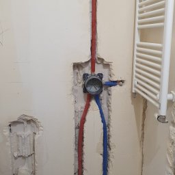 Usługi remontowe Krzysztof Dąbrowski - Ściana z instalacją hydrauliczną w trakcie montażu: czerwone i niebieskie rury prowadzone w bruzdach, puszka instalacyjna, grzejnik łazienkowy na ścianie. Etap prac instalacyjnych.