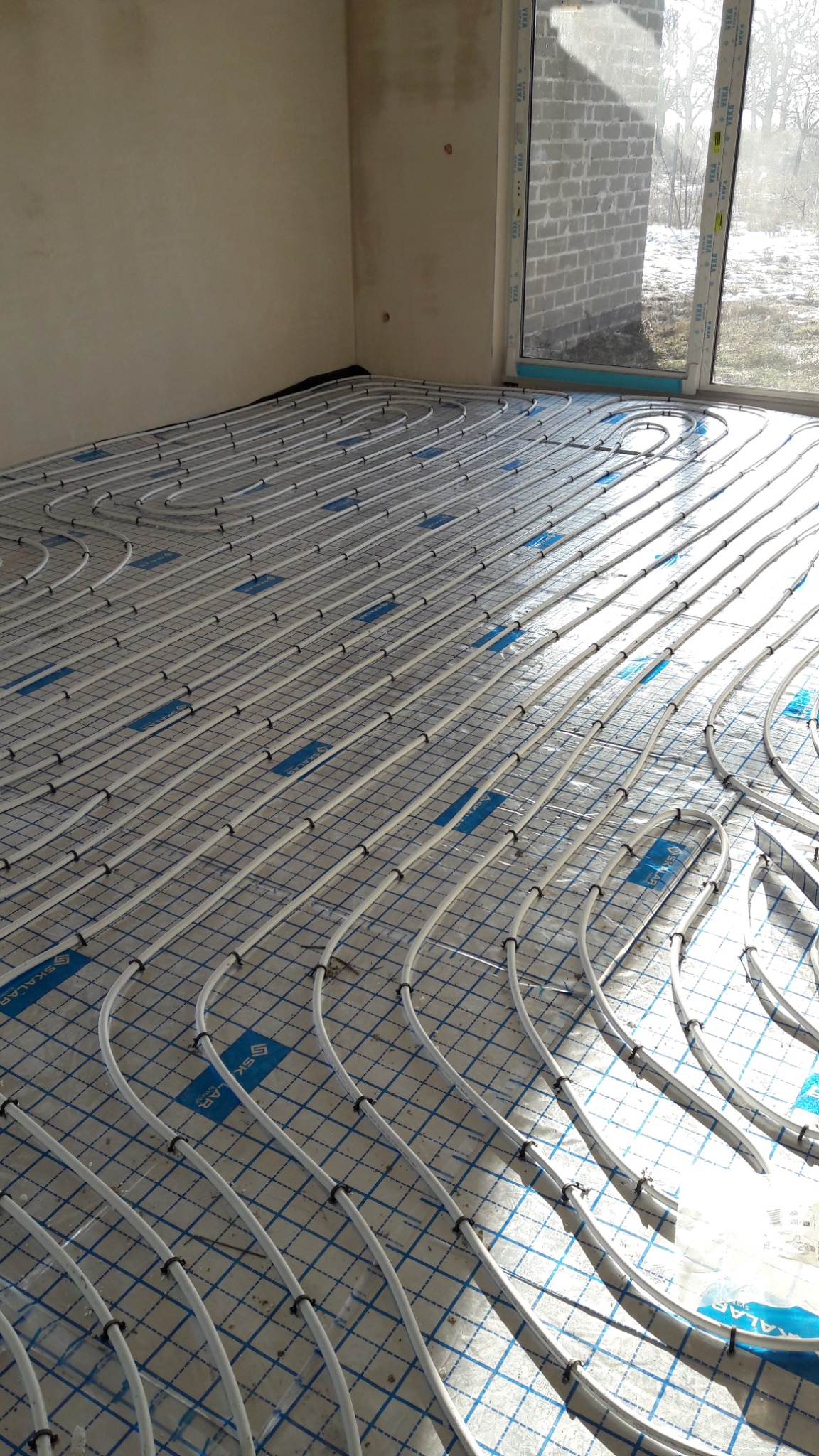 Białe rury ogrzewania podłogowego ułożone równolegle na niebieskiej siatce w jasnym pomieszczeniu z dużym oknem. Widoczny fragment ściany i surowy stan wykończenia.