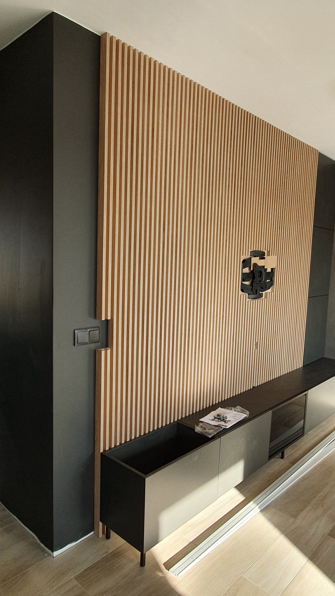 Ściana z drewnianych lameli w odcieniach beżu i brązu, z zamontowanym uchwytem TV, uzupełniona o czarne elementy zabudowy meblowej. Nowoczesne wykończenie wnętrza.