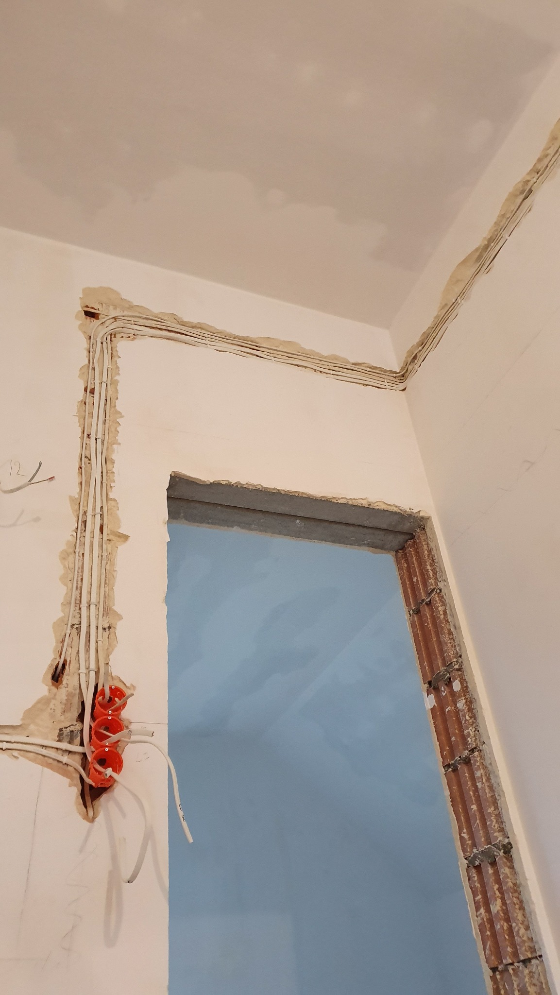 Instalacja elektryczna w bruzdach na ścianie, przed tynkowaniem. Widoczne pomarańczowe puszki i kable. Przygotowanie do remontu, surowy stan, widok z dołu.