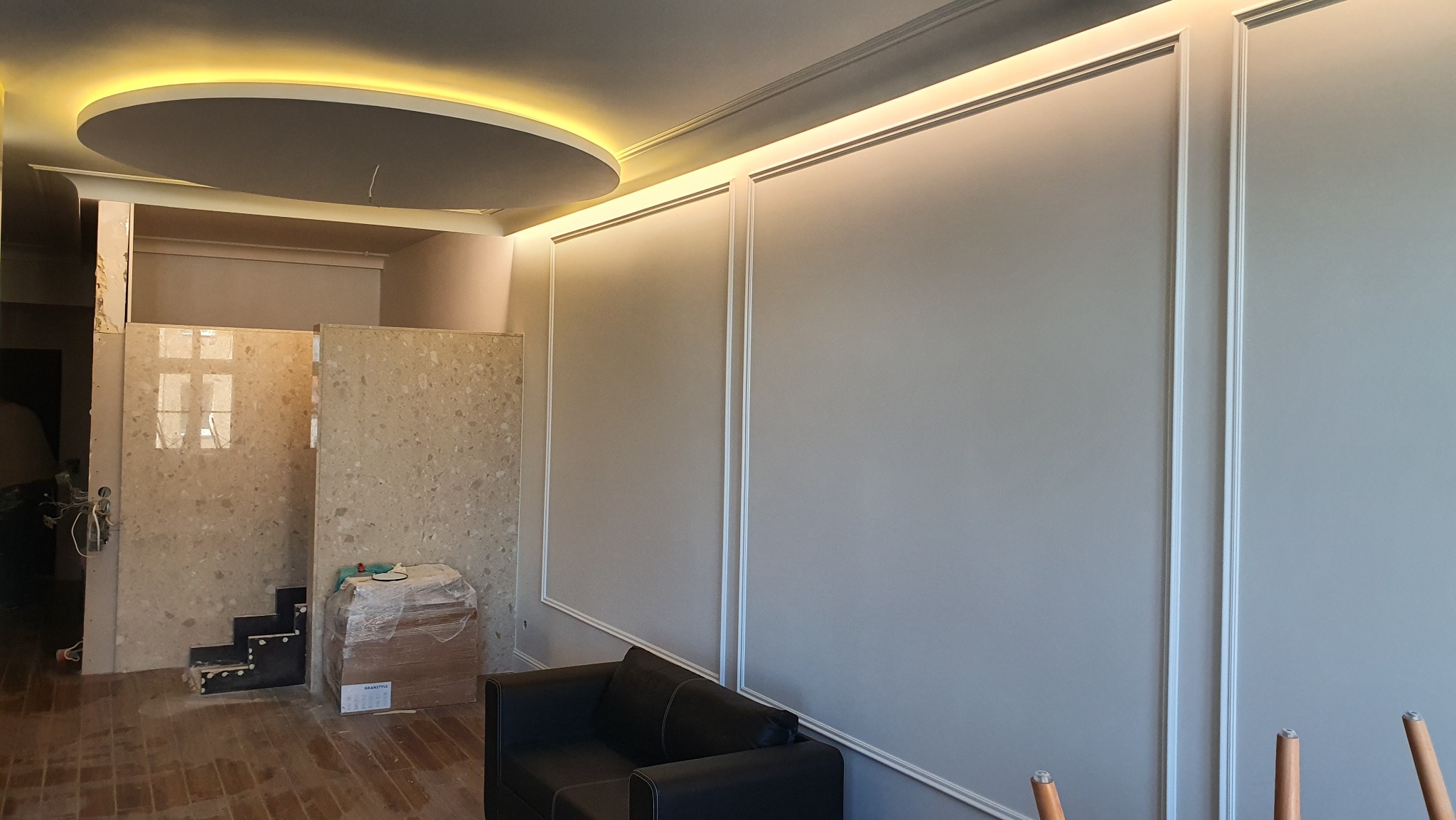 Szara ściana z białymi listwami dekoracyjnymi i oświetleniem LED, widoczny fragment sofy i elementy w trakcie wykończenia, sufit z okrągłym panelem.