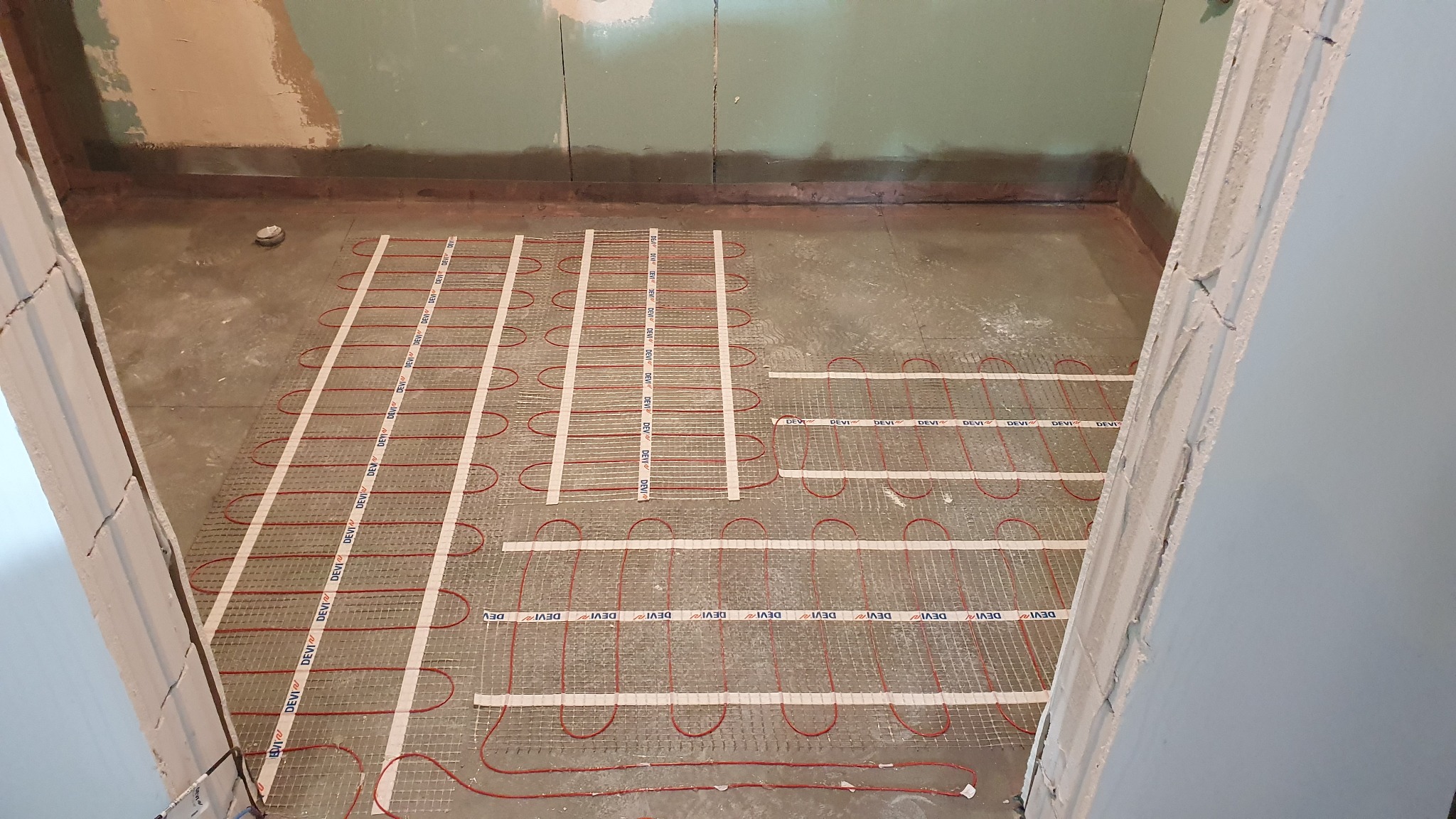 Montaż ogrzewania podłogowego w łazience, widoczne kable grzewcze marki Devi na siatce, przygotowane do zalania wylewką. Ściany surowe, w tle płyta karton-gips.
