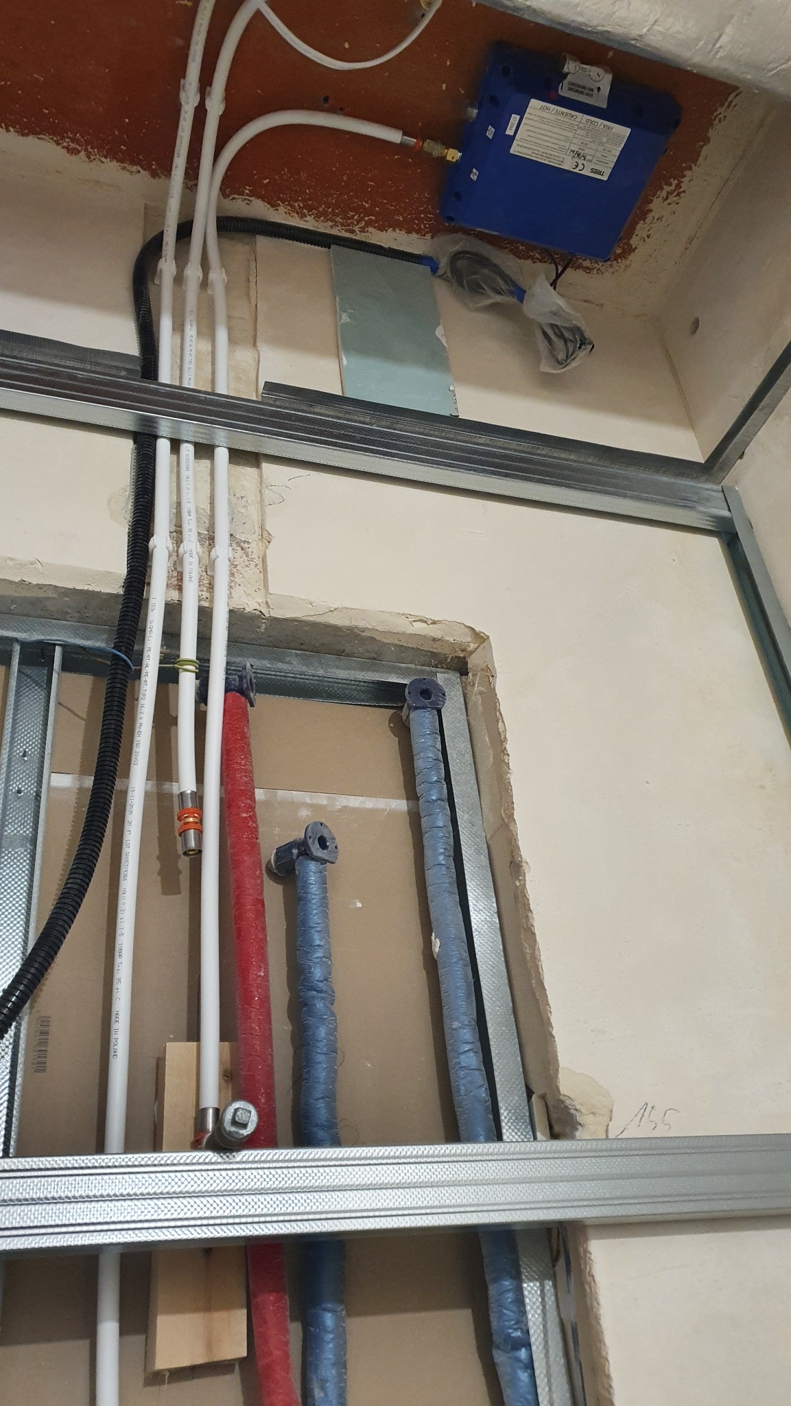 Instalacja wodna w trakcie montażu: białe, czerwone i niebieskie rury prowadzone wzdłuż ściany z profilem metalowym. Widoczny bojler i elementy mocujące.