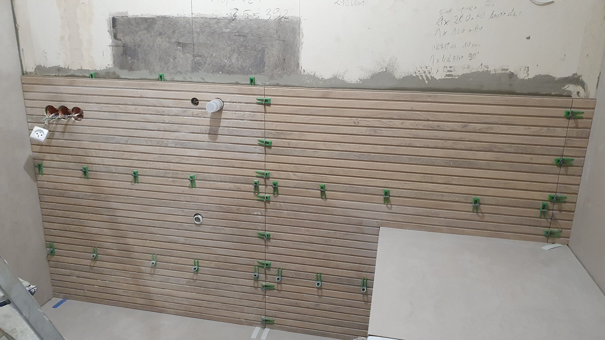 Układanie drewnopodobnej glazury na ścianie z użyciem klipsów poziomujących. Widoczne otwory na instalacje, surowe wykończenie górnej części ściany i notatki.