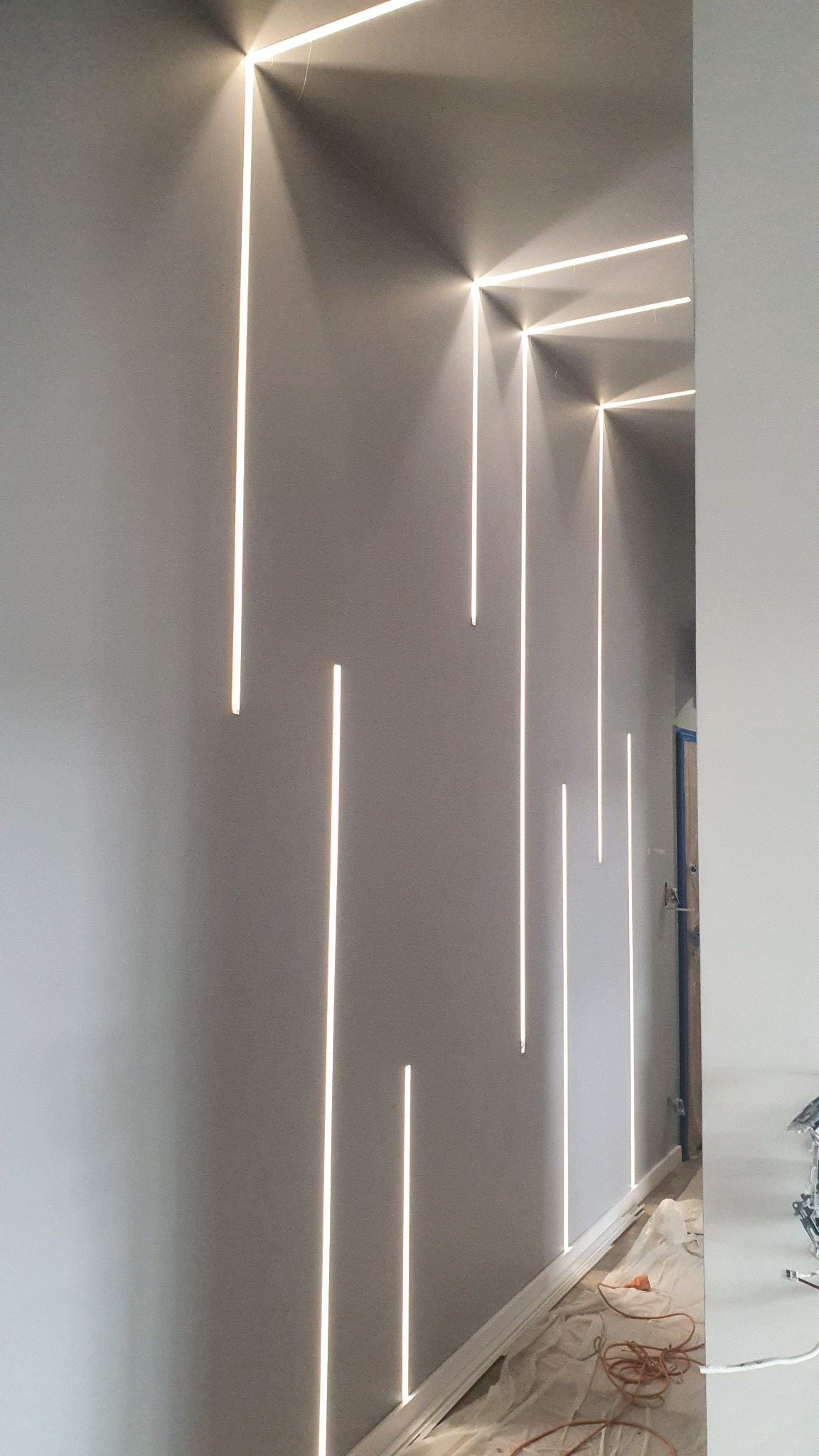 Szara ściana z pionowymi, wpuszczanymi liniami LED, tworzącymi nowoczesny efekt oświetleniowy wzdłuż korytarza. Widoczne zabezpieczenia podłogi podczas prac.