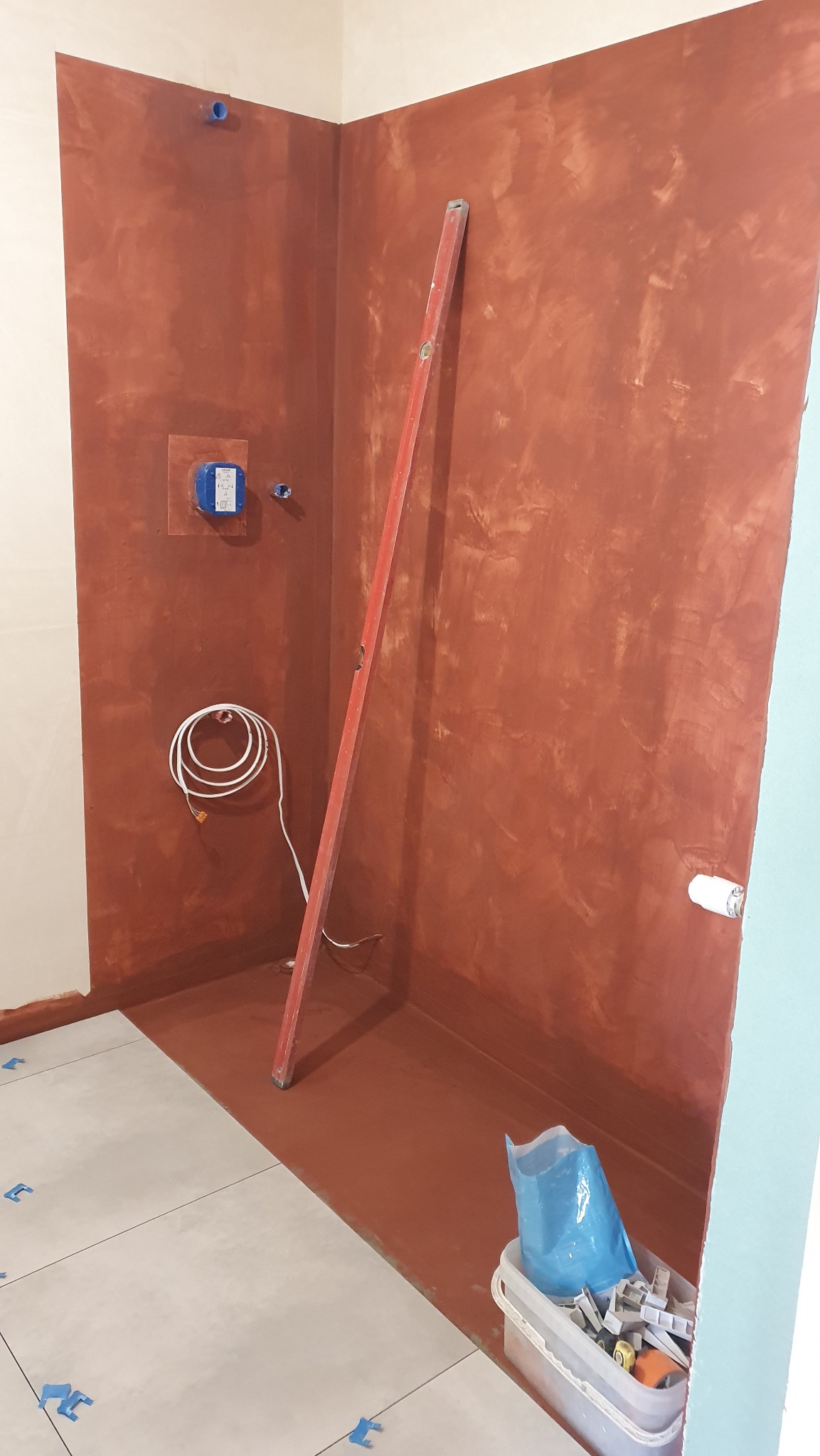 Przygotowanie do wykończenia łazienki: ściany pomalowane na brązowo, poziomica oparta o ścianę, widoczne instalacje, fragment ułożonych płytek podłogowych.