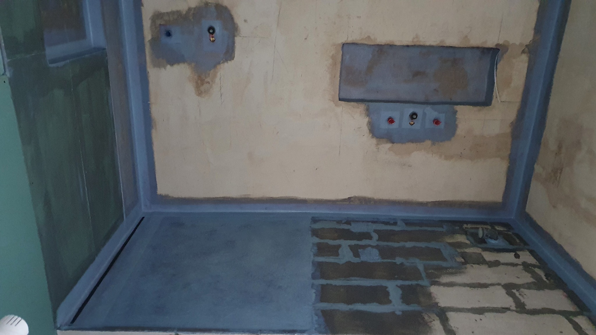 Etap prac w łazience: hydroizolacja ścian i podłogi w szarym kolorze, przygotowanie do dalszych prac wykończeniowych. Widoczne fragmenty instalacji.