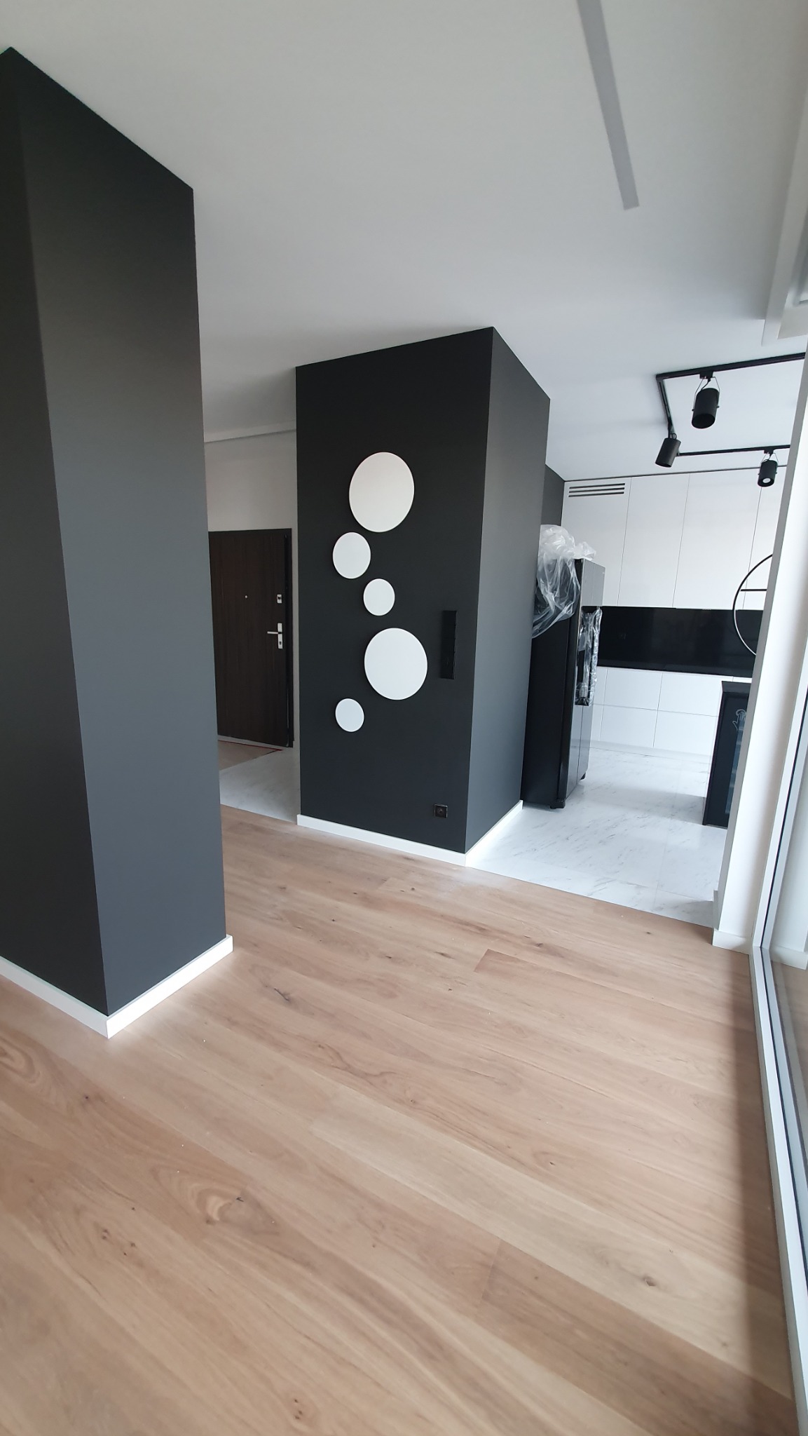 Nowoczesne wykończenie wnętrza: drewniana podłoga, czarne ściany z białymi okręgami, biała kuchnia w tle. Minimalizm i elegancja w aranżacji.