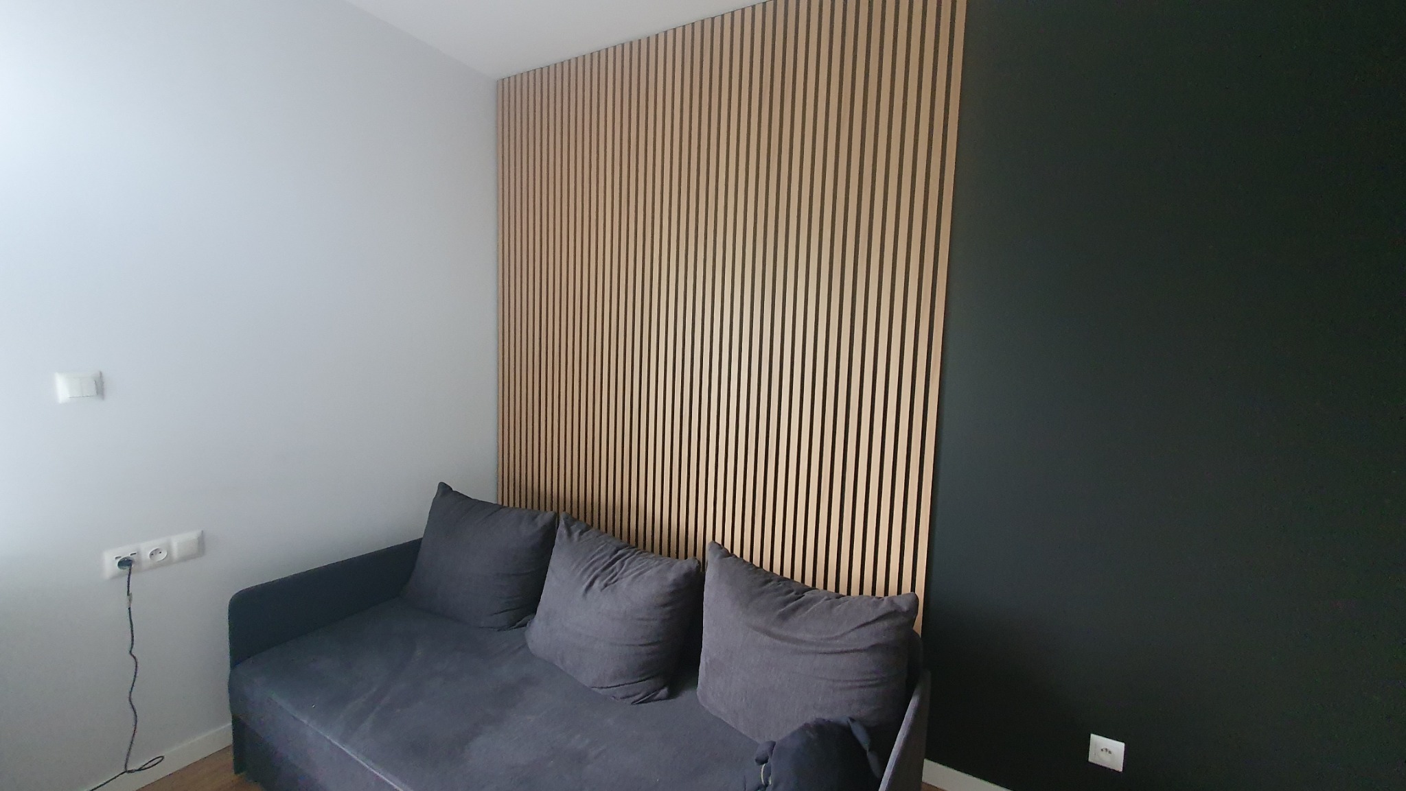 Nowoczesny salon z dekoracyjną ścianą z drewnianych paneli, szarą sofą i ścianami w odcieniach szarości i czerni, tworzący stylowe wnętrze.