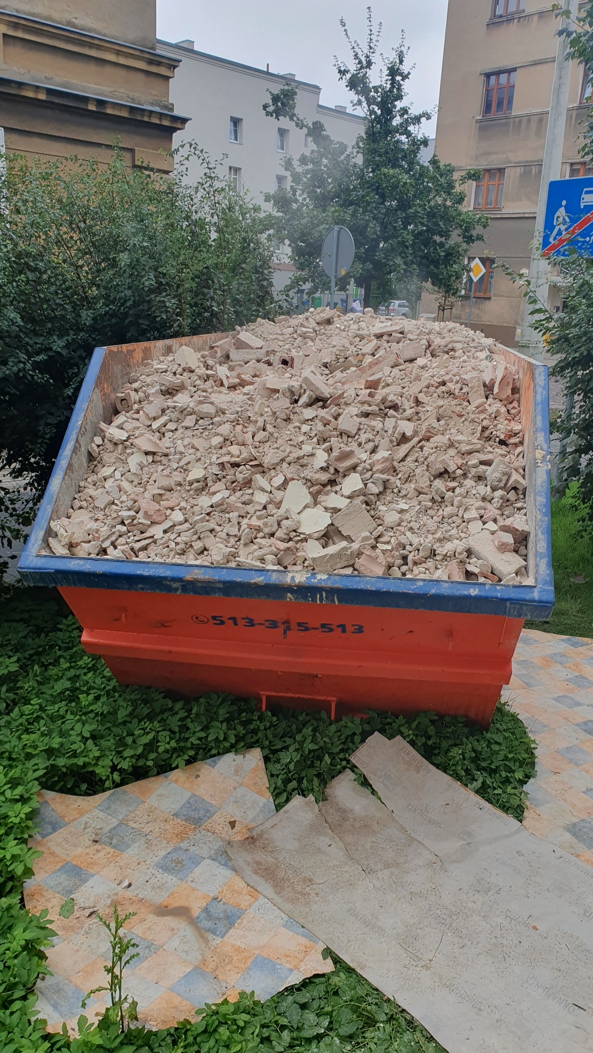Kontener na gruz wypełniony po brzegi, stojący na tle zieleni i budynków miejskich. Widoczne fragmenty potłuczonych płytek i cegieł wewnątrz kontenera.