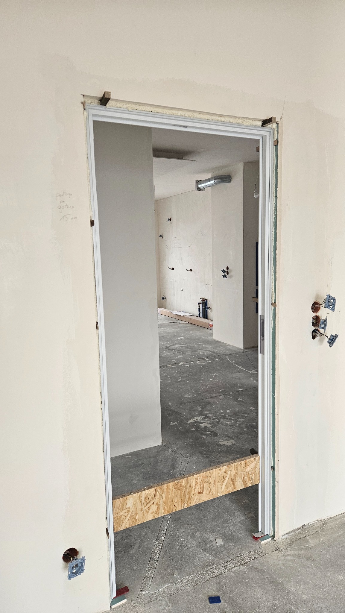 Montaż ościeżnicy w surowym wnętrzu: widoczne przewody, ściany przygotowane do malowania, betonowa podłoga. Etap wykończeniowy z drewnianymi elementami wsparcia.