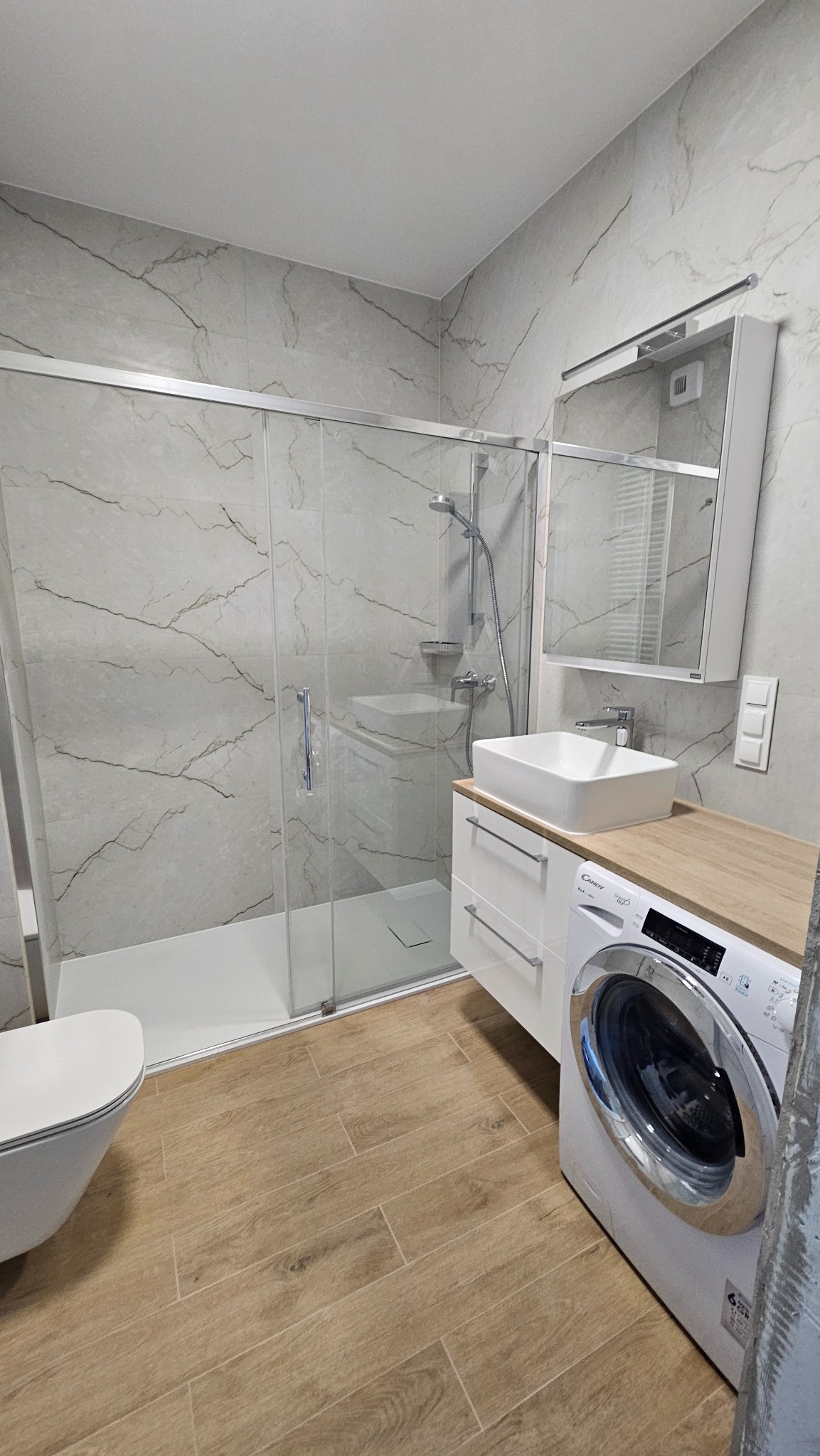 Wykończona łazienka z kabiną prysznicową, umywalką na szafce z drewnianym blatem i pralką. Ściany wyłożone płytkami imitującymi marmur, podłoga drewnopodobna.