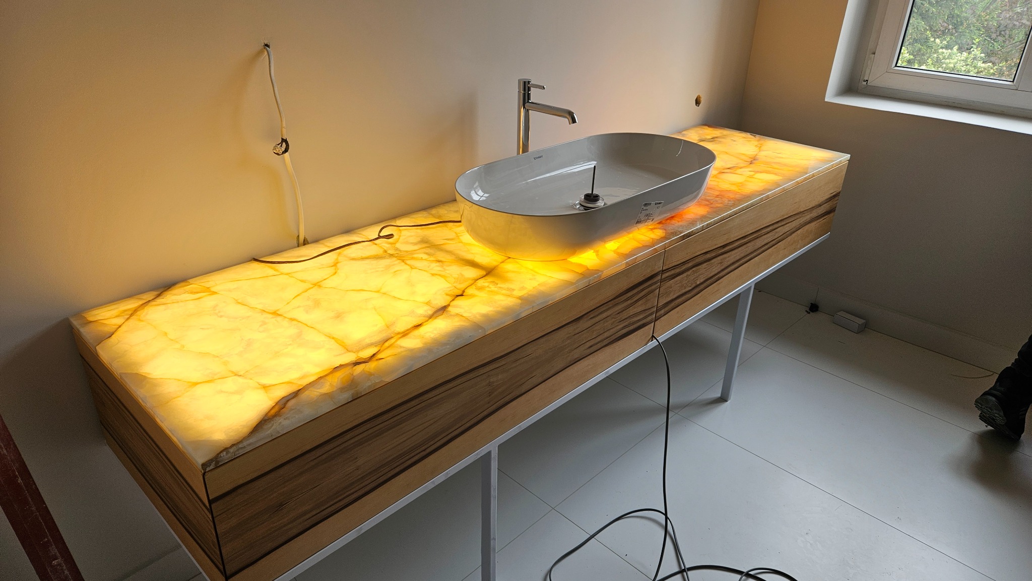 Nowoczesna łazienka: podświetlana szafka z blatem z onyksu i umywalką nablatową. Widoczne srebrne nóżki i fragment instalacji elektrycznej na ścianie.
