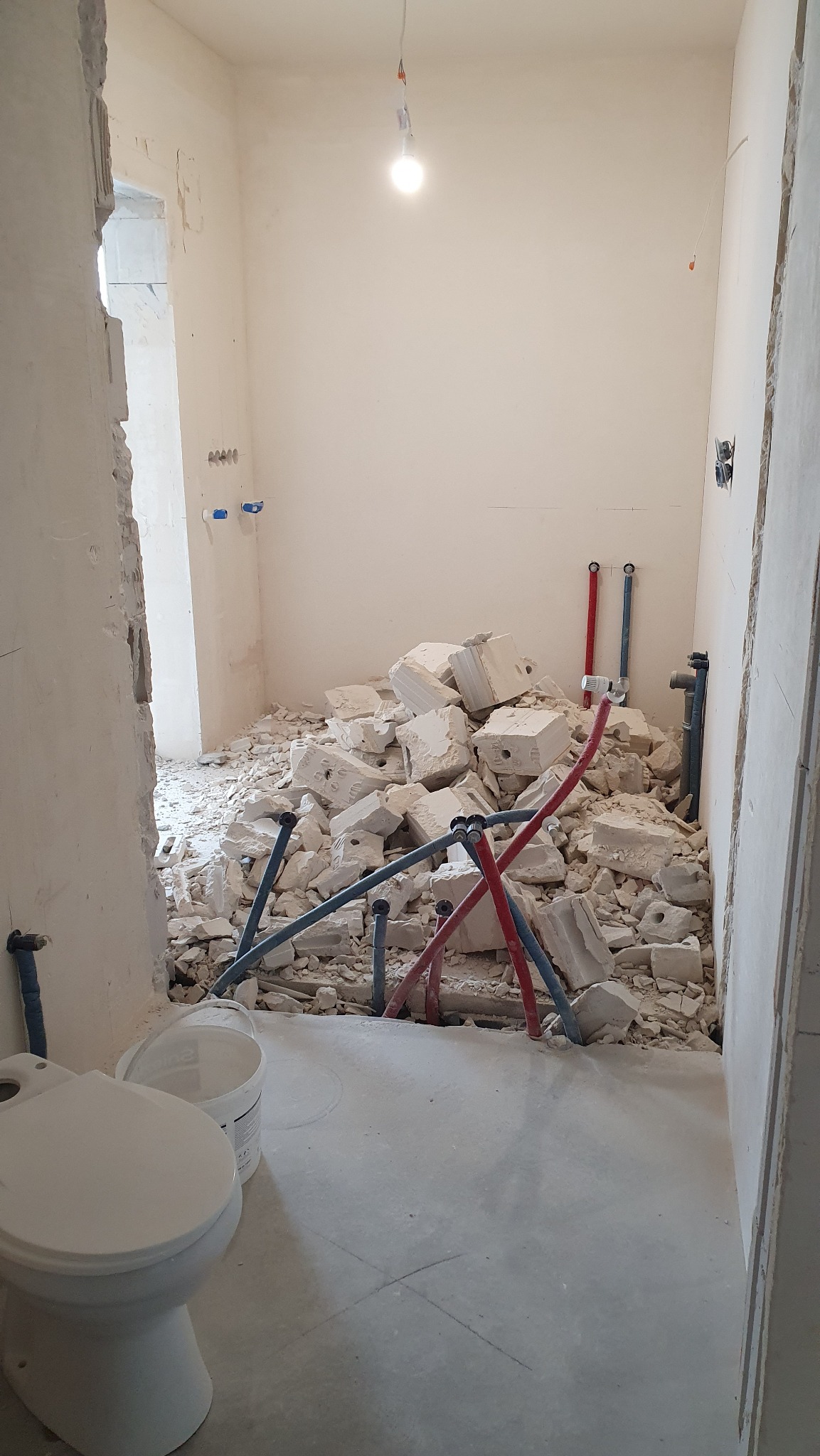 Łazienka w trakcie remontu: gruz po skuciu płytek, wystające rury instalacji wodnej, widoczna toaleta i wiadro. Prace przygotowawcze przed wykończeniem.