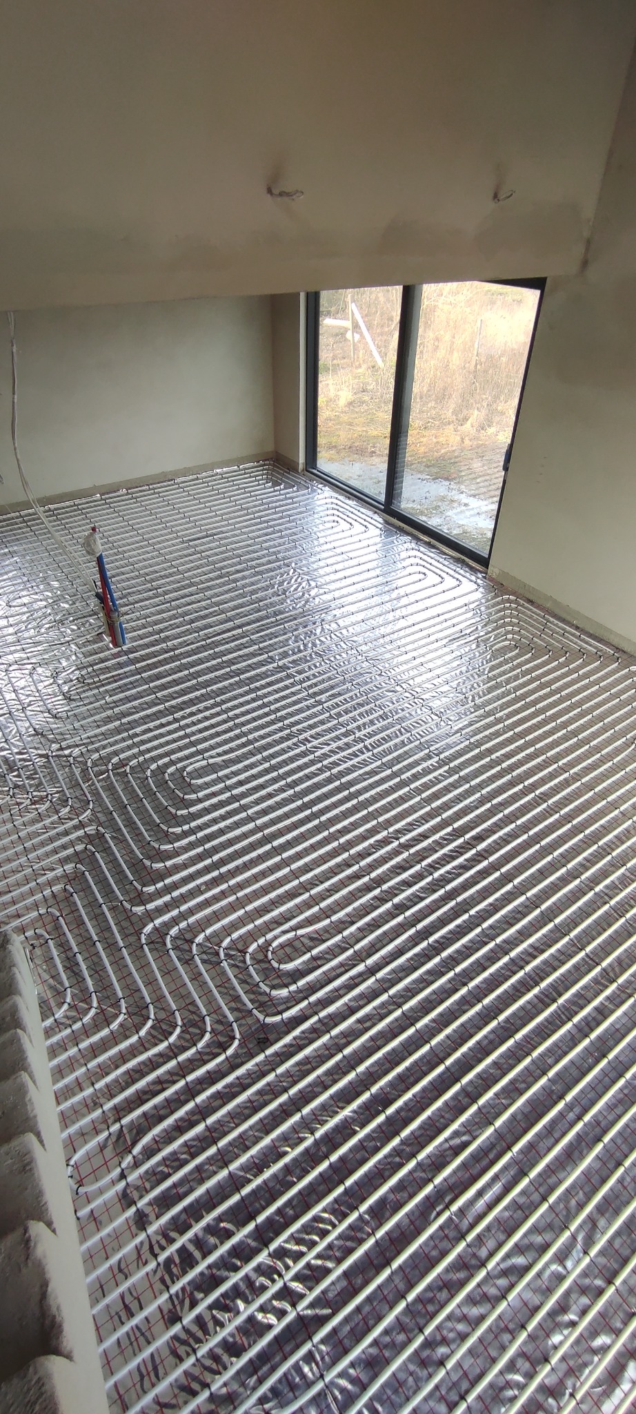 Wnętrze w trakcie montażu ogrzewania podłogowego: rury grzewcze ułożone na folii izolacyjnej z widocznym rozdzielaczem i oknem w tle.