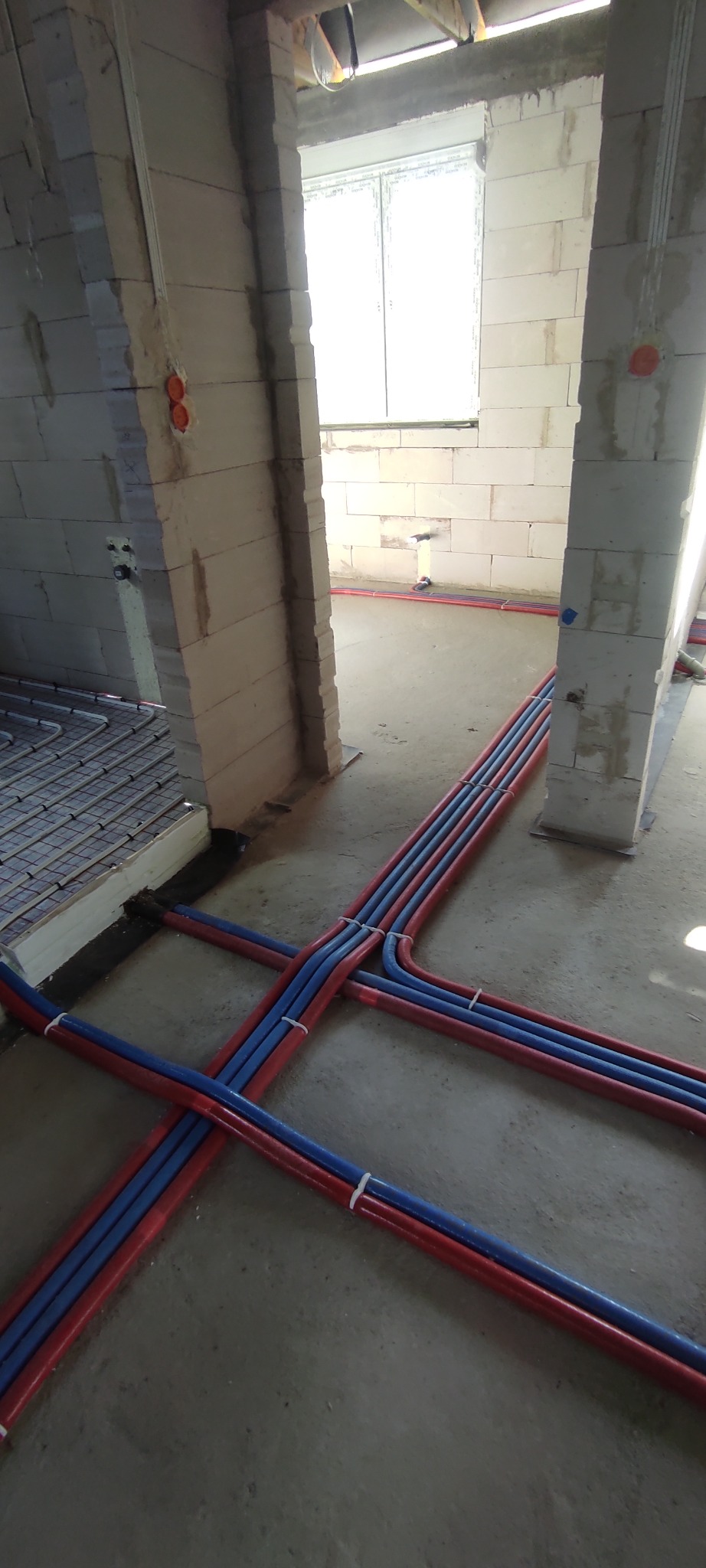 Wnętrze w trakcie budowy z widocznymi rurami ogrzewania podłogowego oraz instalacji wodnej w czerwono-niebieskiej osłonie, przymocowanymi opaskami zaciskowymi do podłoża, z fragmentem rozłożonej...