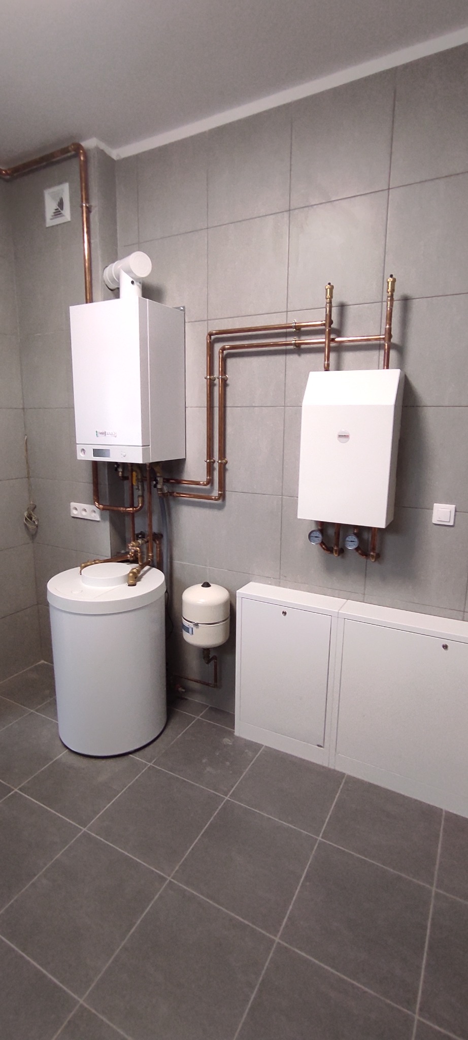 Biały piec centralnego ogrzewania z bojlerem i rurami miedzianymi na tle szarych płytek w pomieszczeniu technicznym.