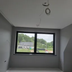 Świeżo pomalowane wnętrze pokoju z szarymi ścianami, czarną ramą okienną z widokiem na dom i zieleń, oraz wystającymi przewodami elektrycznymi z sufitu.