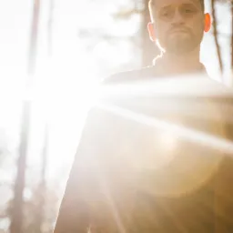 Portret mężczyzny z brodą w lesie, oświetlony silnym światłem słonecznym, z widocznymi promieniami przebijającymi się przez drzewa.