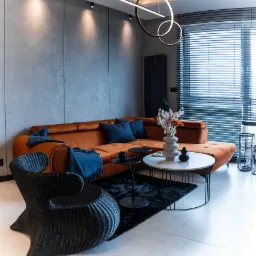 Nowoczesny salon z pomarańczową sofą narożną, ciemnoniebieskimi poduszkami i pledem, czarnym fotelem z rattanu, okrągłym stolikiem kawowym z białym blatem, czarnym dywanem i designerską lampą sufitową