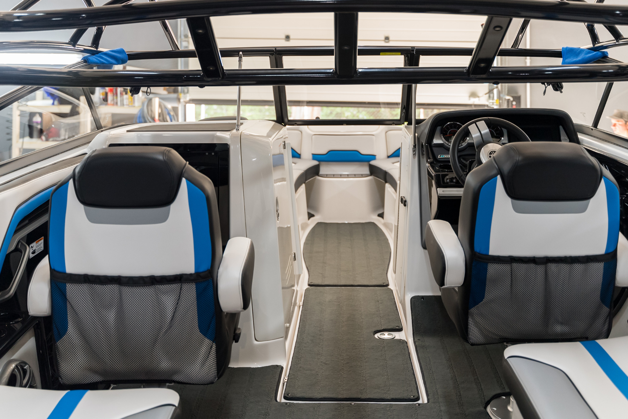 Wnętrze nowoczesnej łodzi motorowej z białymi ścianami, czarno-biało-niebieskimi siedzeniami i czarną ramą nad głową, widok z tyłu na kabinę sterowniczą i otwartą przestrzeń na zewnątrz.