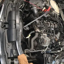 Otwarta maska samochodu BMW E90 z widocznym silnikiem podczas naprawy układu paliwowego. Widoczne wtryskiwacze, przewody paliwowe i inne elementy silnika. Na pierwszym planie fragment brudnej...