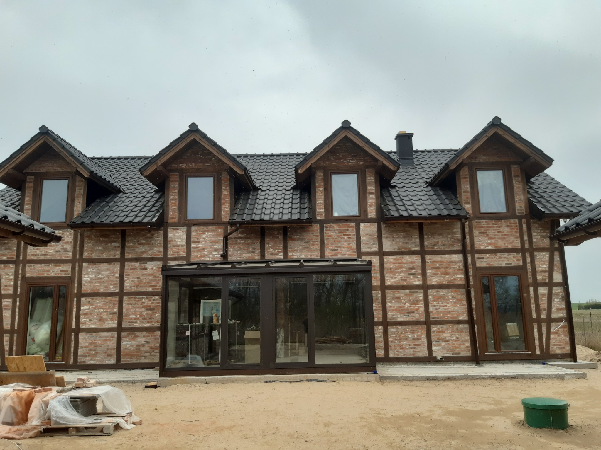 Dwukondygnacyjny dom murowany w stylu szachulcowym z ciemną dachówką, widoczna przeszklona weranda i elementy wykończeniowe na zewnątrz.