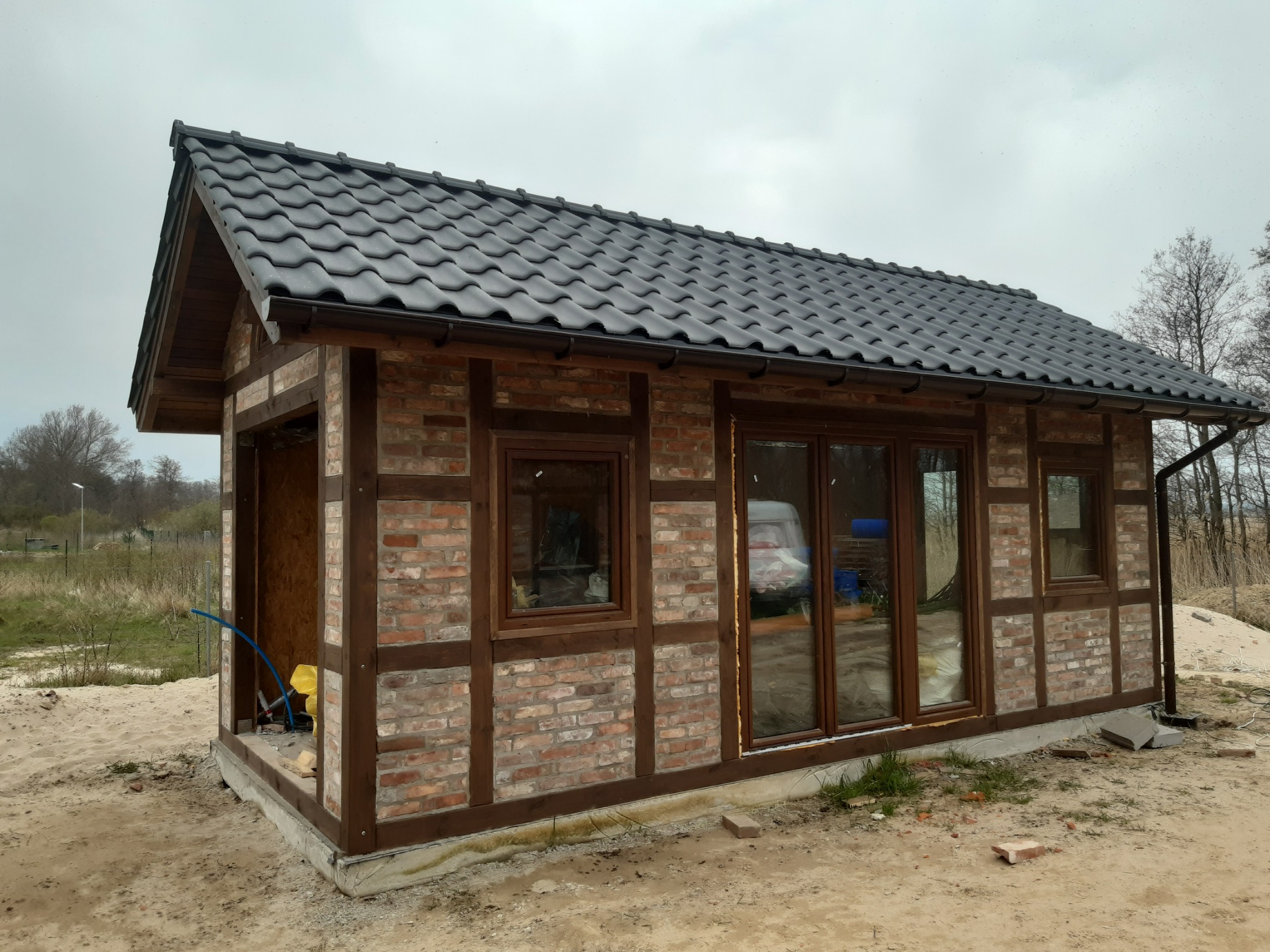 Niedokończony budynek z cegły klinkierowej z ciemnymi, drewnianymi belkami konstrukcyjnymi, czarną dachówką i widocznymi instalacjami wewnątrz, otoczony piaszczystym terenem budowy.