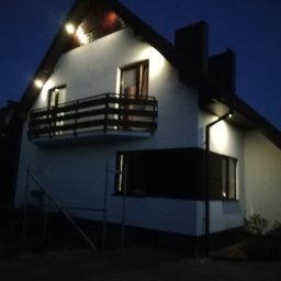 Przemek Suchocki - Dom z balkonem oświetlony zewnętrznymi lampami w nocy, widoczne rusztowanie, elewacja w kolorze białym z ciemnymi detalami.