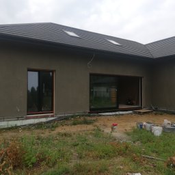 Przemek Suchocki - Surowa elewacja domu jednorodzinnego z ciemną dachówką, widoczne okna z ciemnymi ramami, drabina oparta o ścianę oraz teren budowy z wiadrami i narzędziami.
