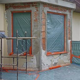 Przemek Suchocki - Przygotowanie elewacji do prac wykończeniowych: okna zabezpieczone folią, ściany z fragmentami starego tynku i świeżym podkładem, widoczne rusztowanie.