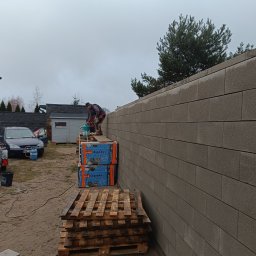 Przemek Suchocki - Wznoszenie ściany z szarych bloczków betonowych; murarz na paletach wyrównuje poziom, w tle samochód osobowy i szara budka narzędziowa.
