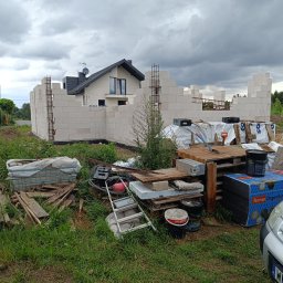 Przemek Suchocki - Budowa domu w toku: wzniesione ściany z białych bloczków, widoczne zbrojenia, w tle gotowy dom. Na pierwszym planie materiały budowlane i narzędzia.