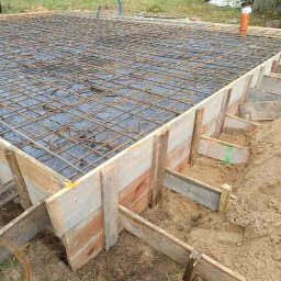 Przemek Suchocki - Zbrojenie fundamentów w drewnianym szalunku, widoczne pręty i folia. Prace ziemne, teren budowy z piaskiem i zielenią w tle. Elementy konstrukcyjne z bliska.