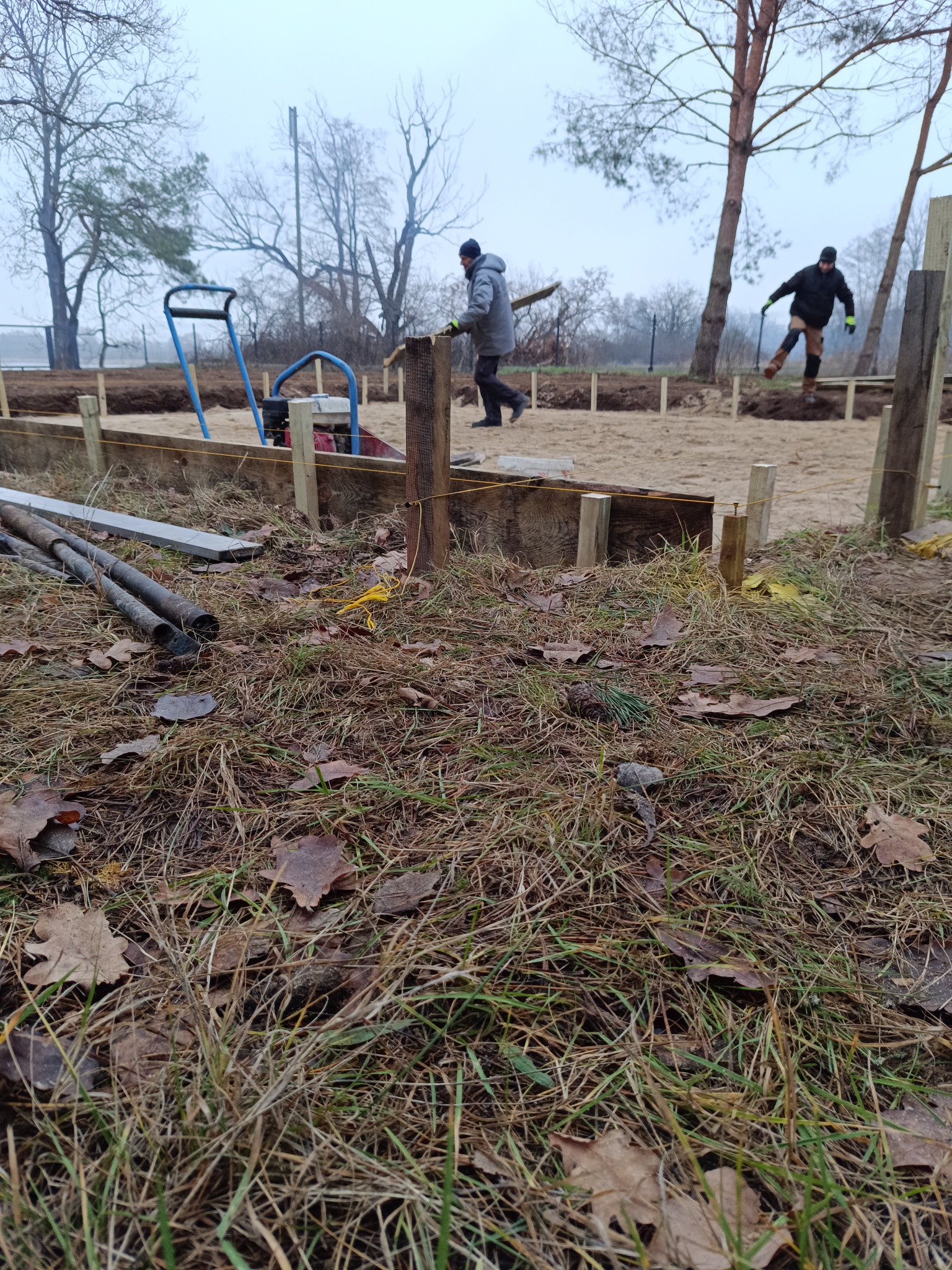 Przygotowanie terenu pod fundamenty: widoczne drewniane szalunki, poziomowanie gruntu, pracownicy w oddali, sprzęt budowlany i rury na pierwszym planie.