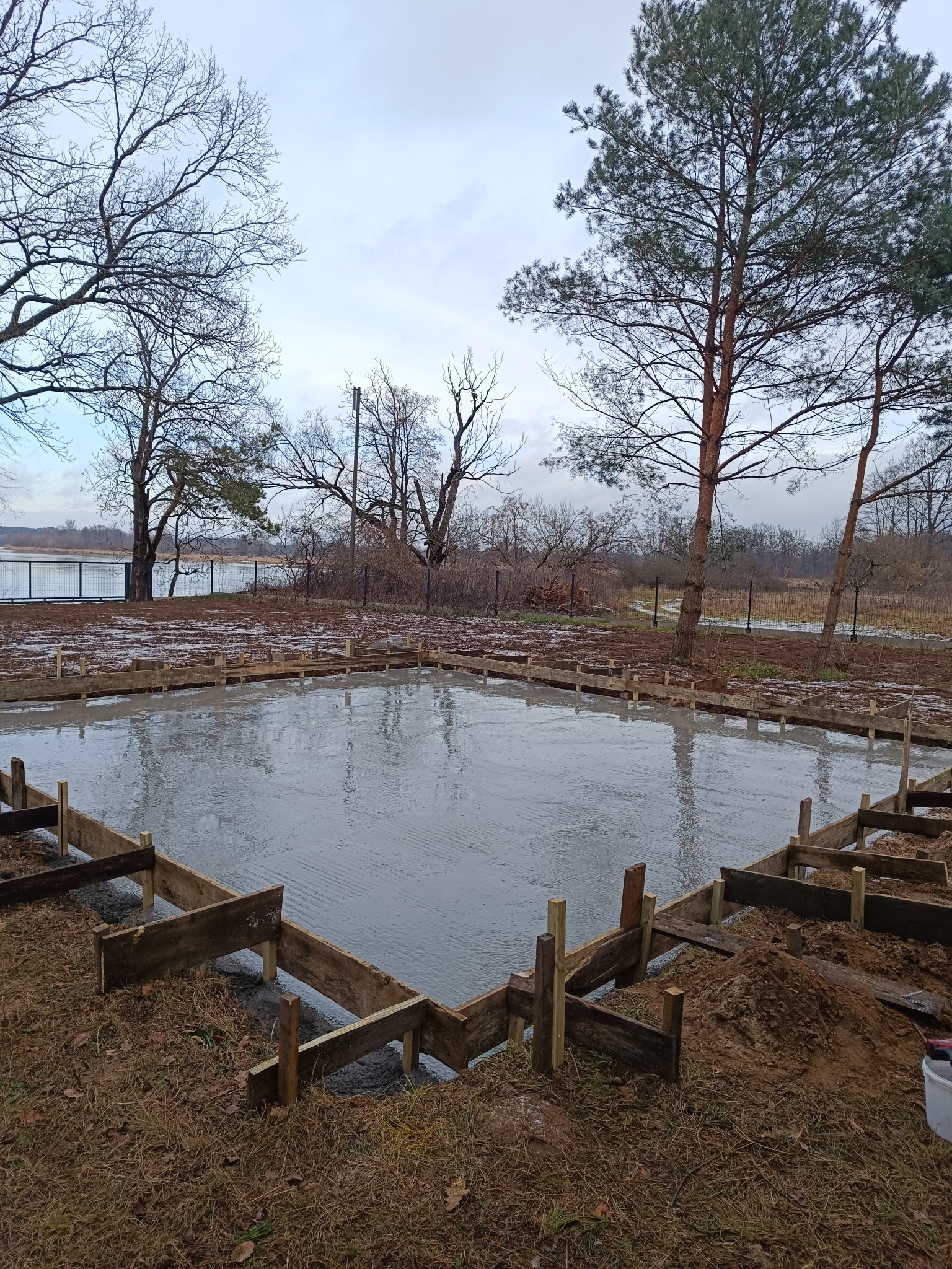 Wylany fundament pod budynek z drewnianym szalunkiem w kształcie prostokąta, widok na jezioro i drzewa w tle.